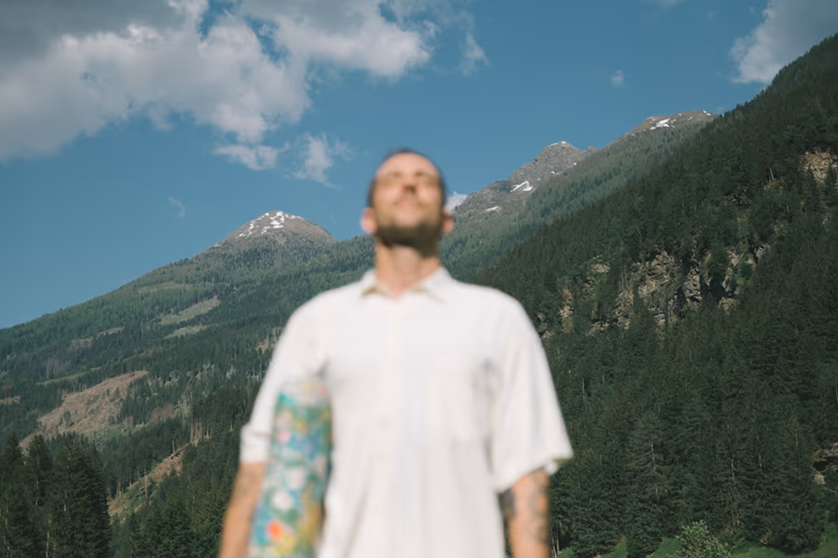 Mann mit einem Skateboard steht vor einer bewaldeten Berglandschaft unter blauem Himmel mit Wolken.