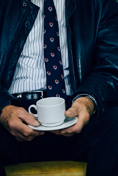 Mann in gestreiftem Hemd, gemusterter Krawatte und Lederjacke hält eine weiße Tasse mit Untertasse in beiden Händen.