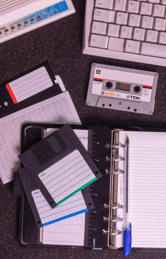 Floppy-Disketten, eine Audiokassette, eine Computertastatur und ein Organizer mit Stift auf schwarzem Tisch.