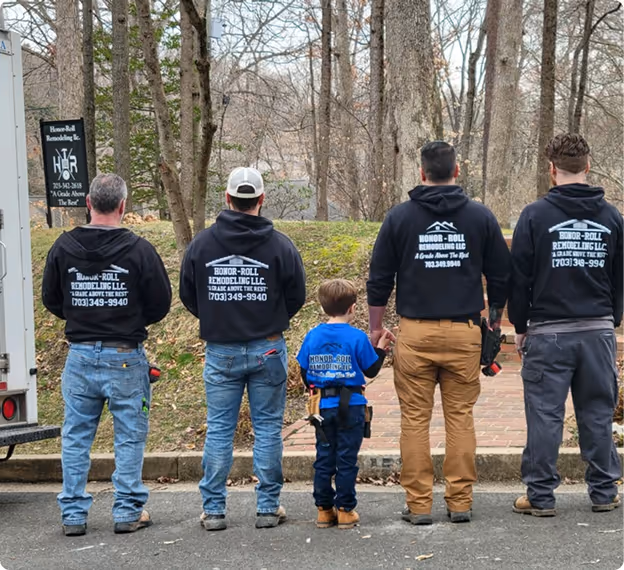 Five people, including a child, standing with their backs to the camera wearing Honor-Roll Remodeling LLC hoodies, outdoors near trees and a brick pathway.