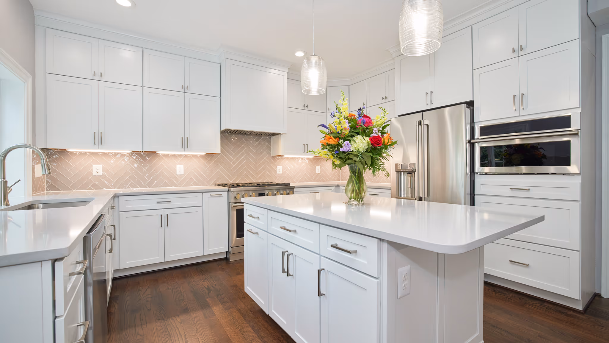 Modern white kitchen with stainless steel appliances, a large island with drawers, and a colorful flower arrangement on the island.