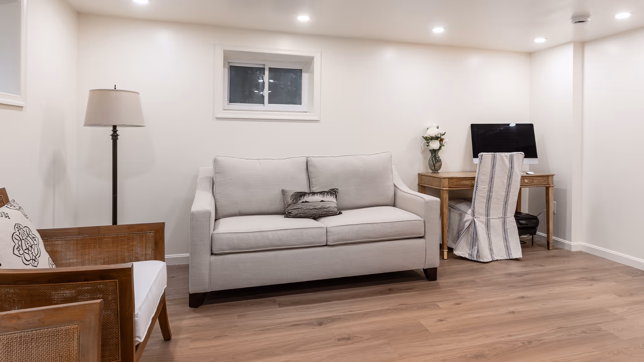 Modern living room with beige sofa, wooden armchair, floor lamp, and a wooden desk with computer and striped chair cover.
