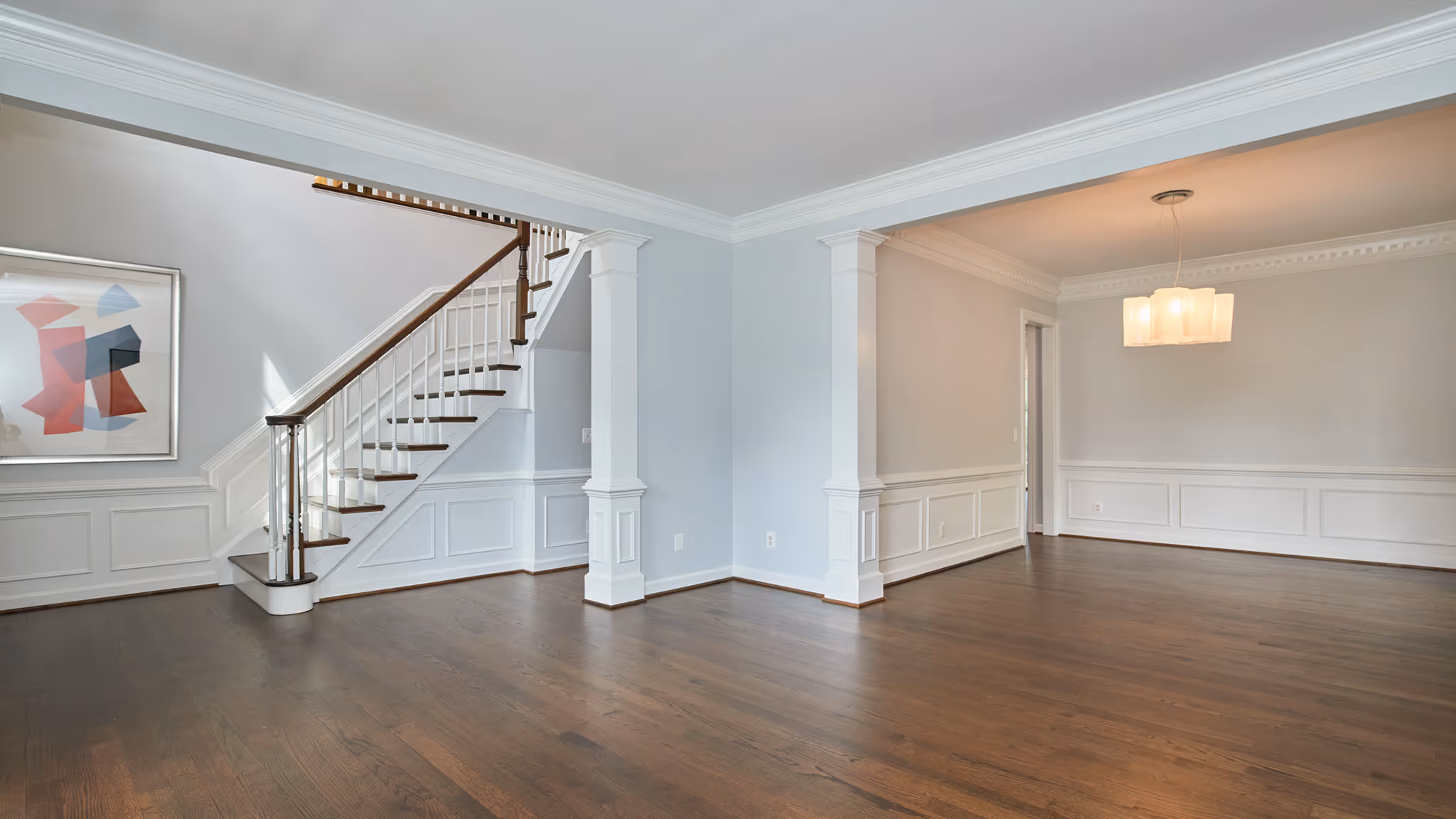 Empty modern living room with dark hardwood floors, white wainscoting, a staircase with wooden handrail, and a hanging light fixture.