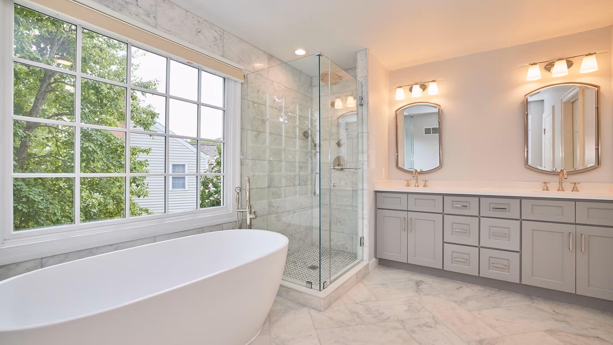 Bright bathroom with large window, freestanding white bathtub, glass-enclosed marble shower, and double vanity with mirrors and lights.