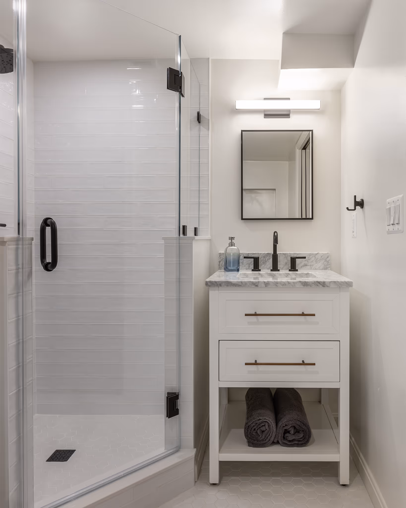 Modern bathroom with white vanity, marble countertop, black fixtures, two rolled black towels below, and a glass shower enclosure with white tile walls.