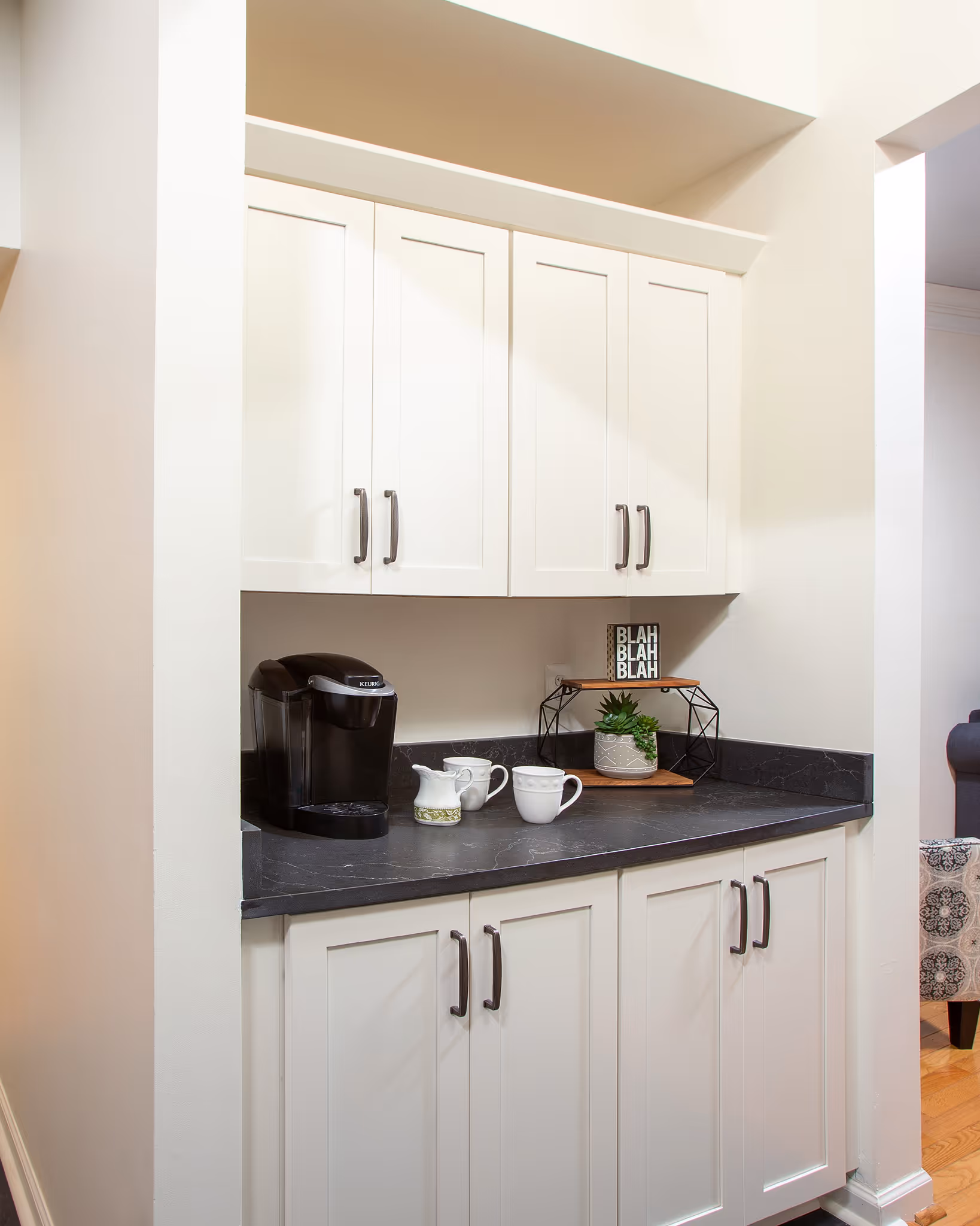 Compact kitchen corner with black countertop, white cabinets, coffee maker, mugs, and a small plant with a decorative sign.