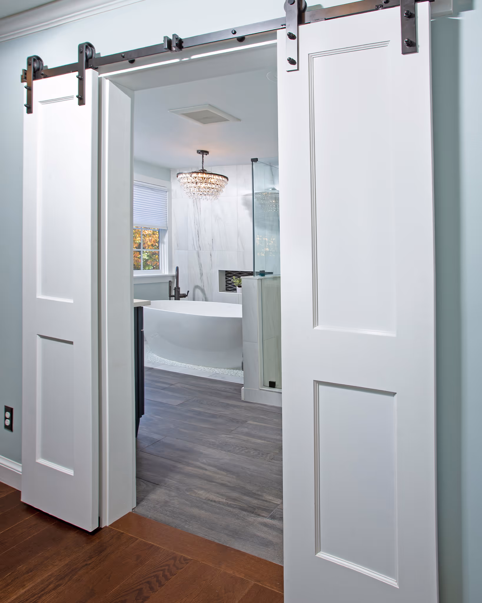 View through open white sliding barn doors into a bathroom with a freestanding bathtub, gray wood-look floor, and a crystal chandelier.