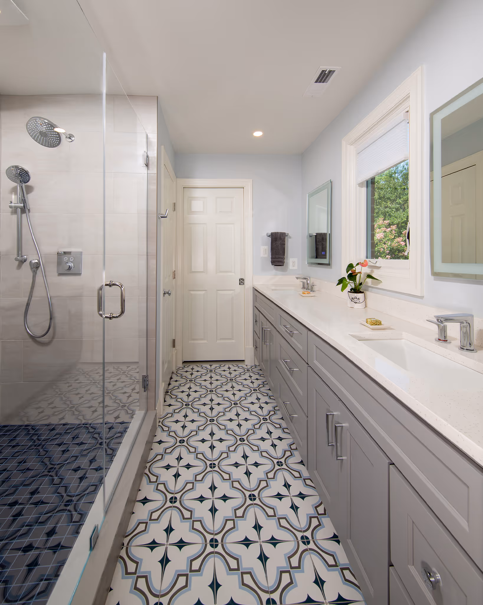 Modern bathroom with patterned floor tiles, gray cabinets with dual sinks, a large mirror, glass shower enclosure, and a window showing green foliage outside.