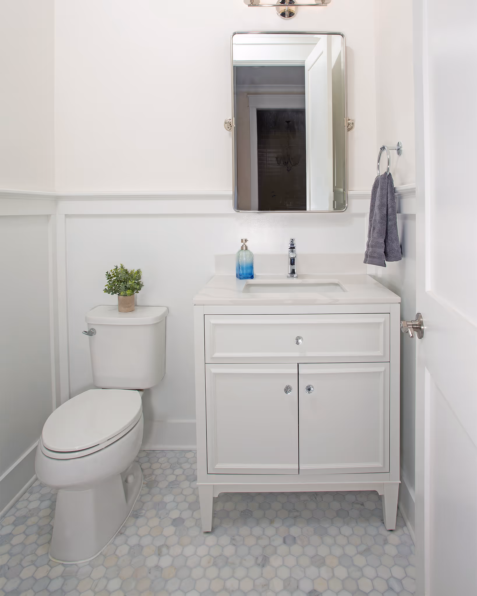 Minimalist white bathroom with a toilet topped by a small plant, a white vanity with a sink, a blue soap dispenser, and a mirror above.