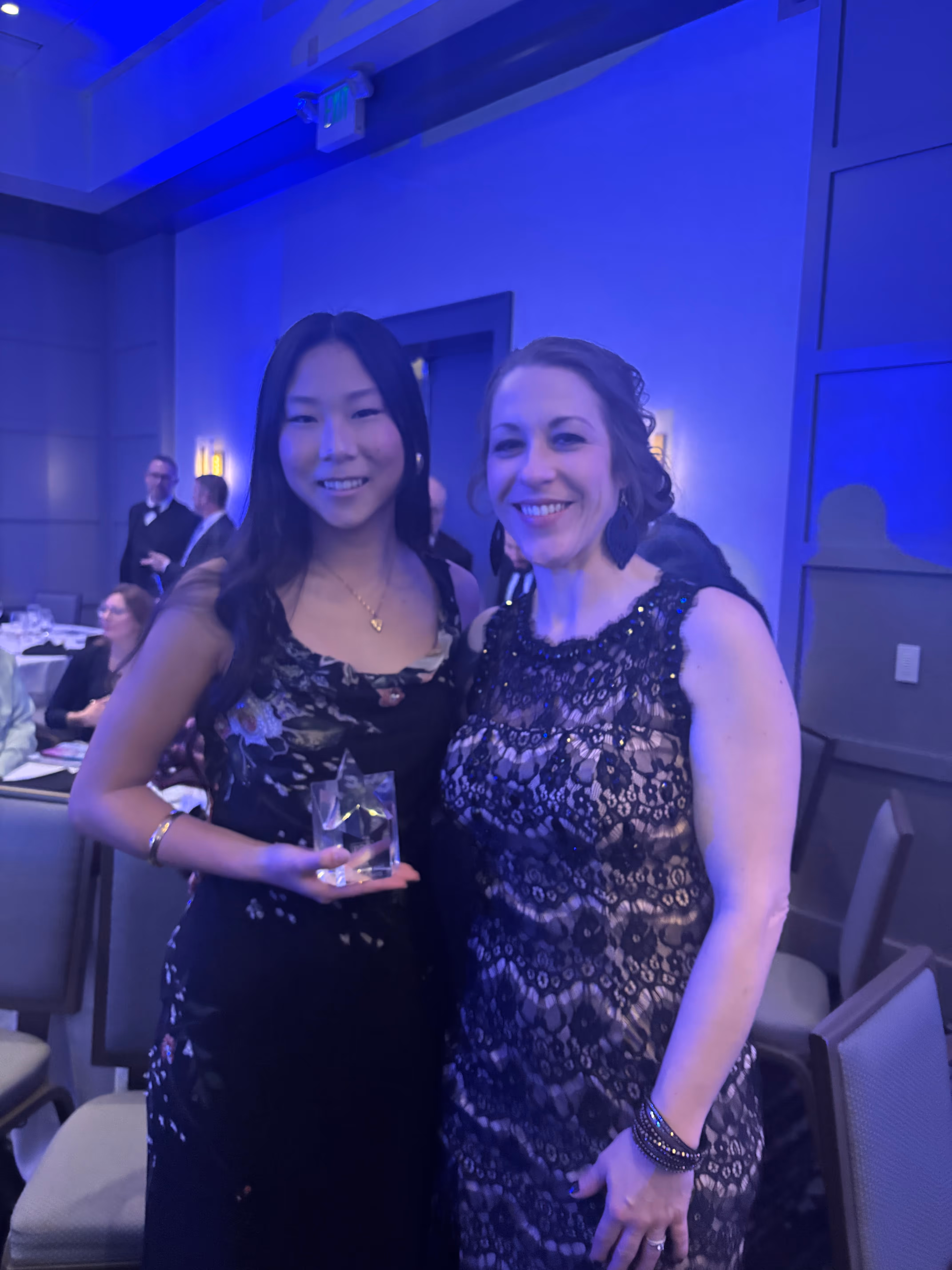 Two women dressed in elegant black dresses smiling; one woman holds a crystal star-shaped award at an indoor event.