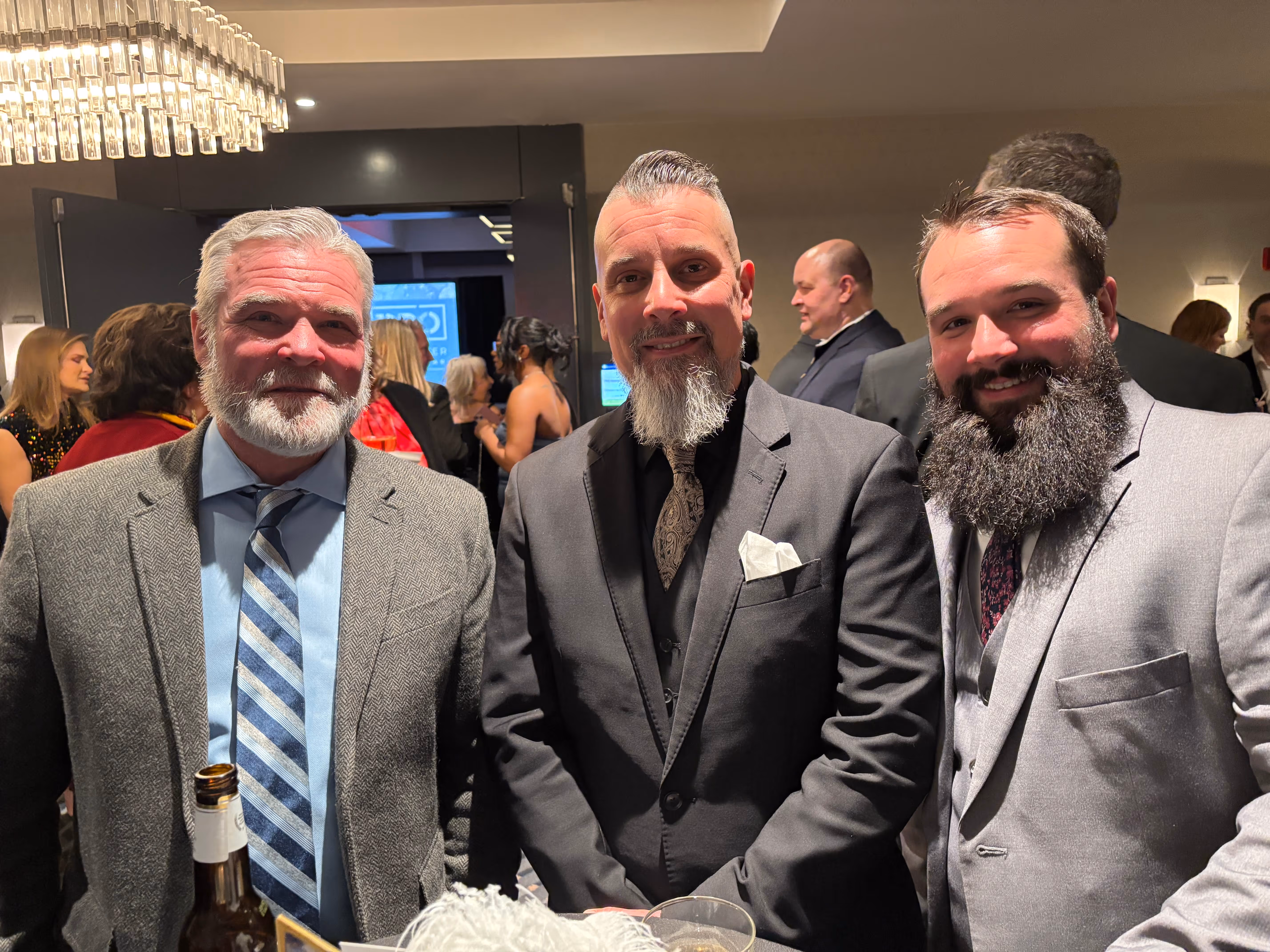 Three men in suits with beards smiling at a formal indoor event.