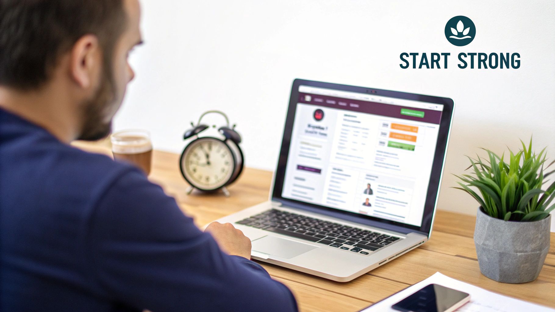 A man with a beard sits at a wooden desk, focused on his laptop screen with a drink.