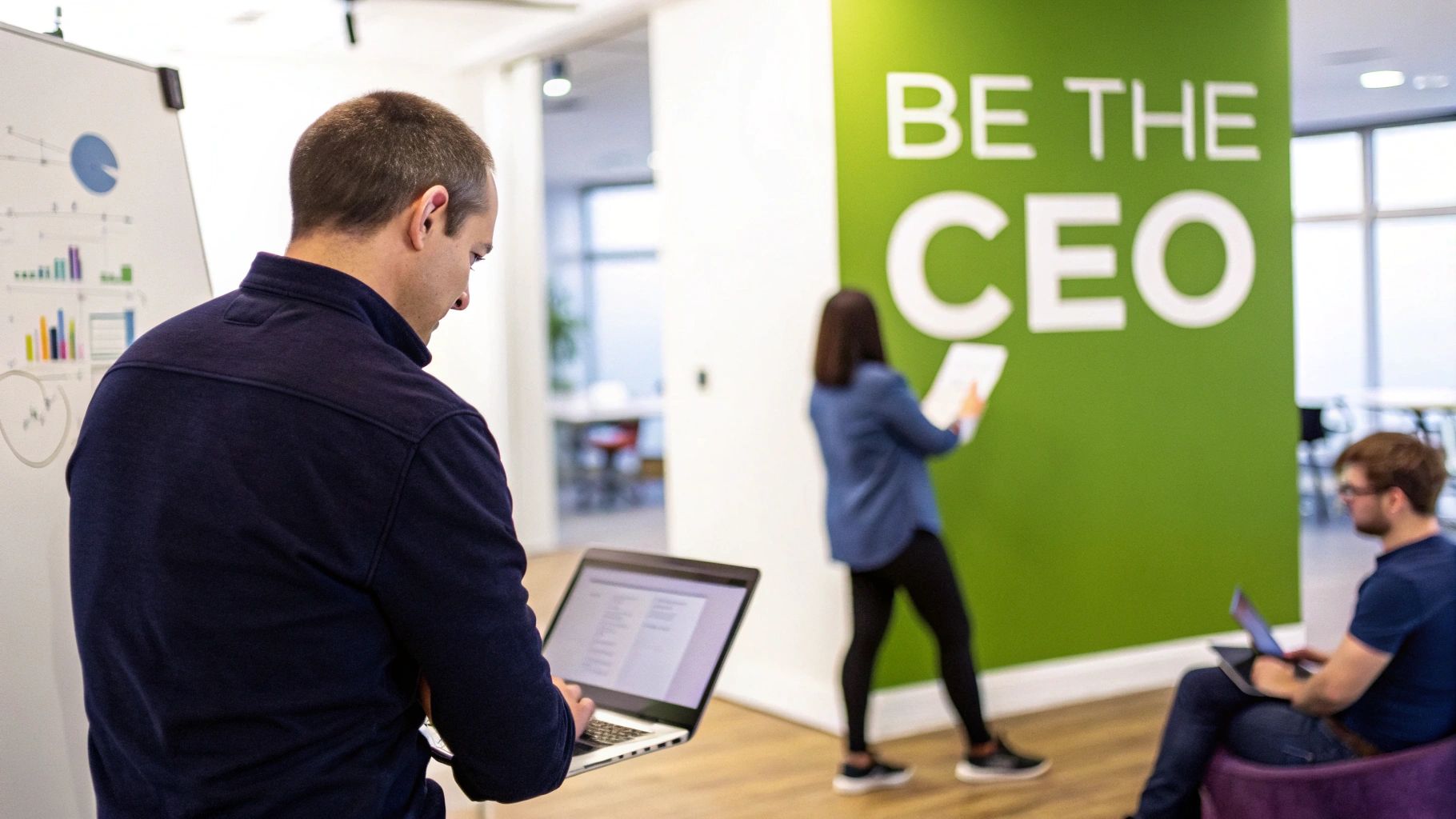 A man on a laptop near a whiteboard, with colleagues in a modern office space.
