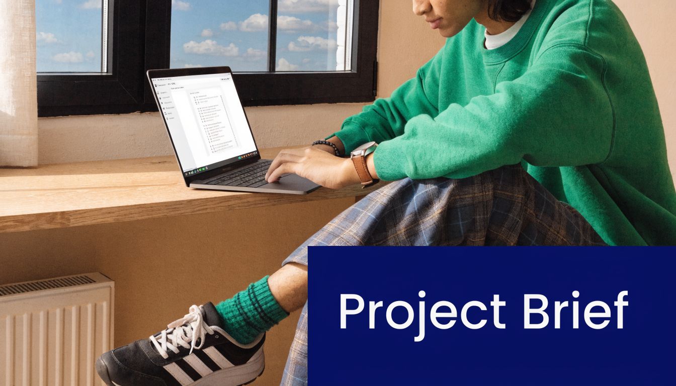 A person wearing a green sweater sits by a window while working on a project brief on their laptop.
