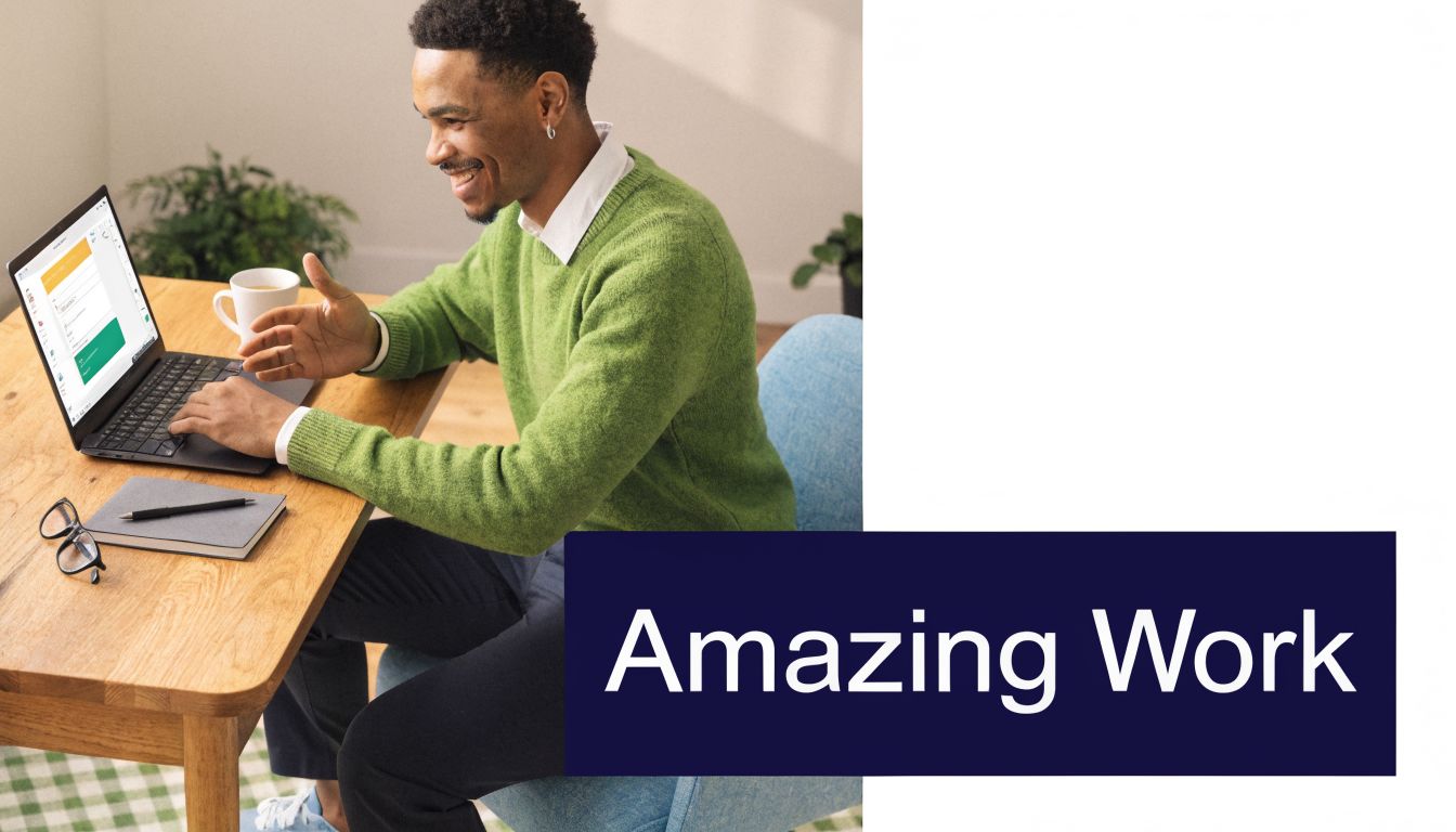 A smiling professional in a green sweater working on a laptop at a wooden desk.