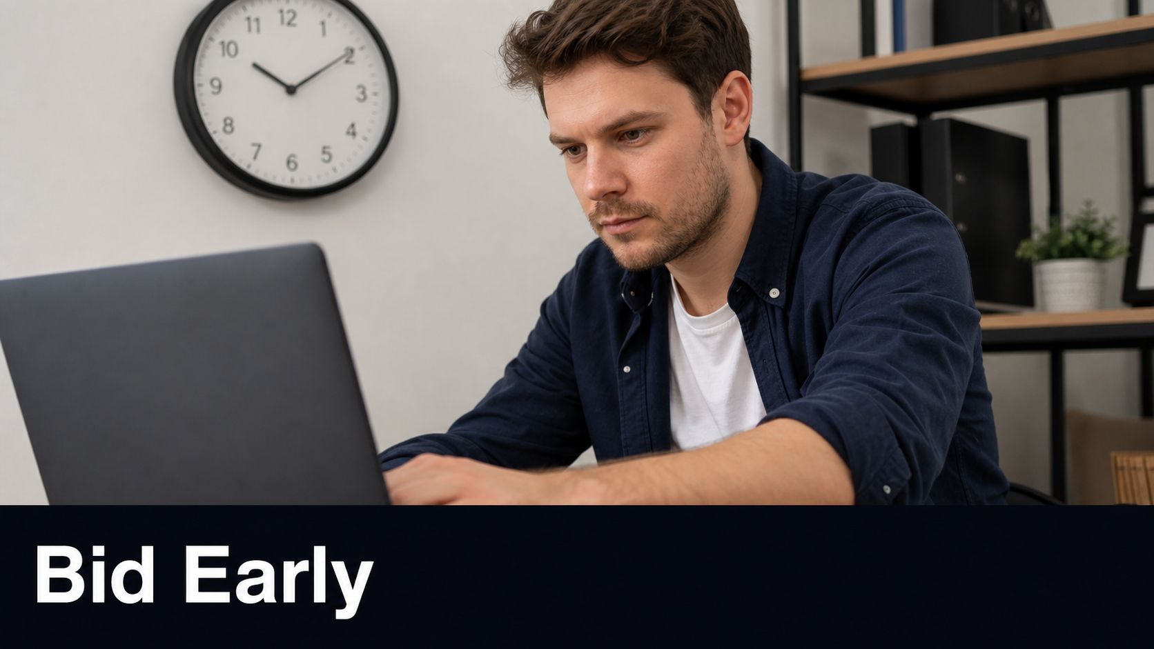 A focused man working on his laptop while sitting at a desk with a wall clock nearby.
