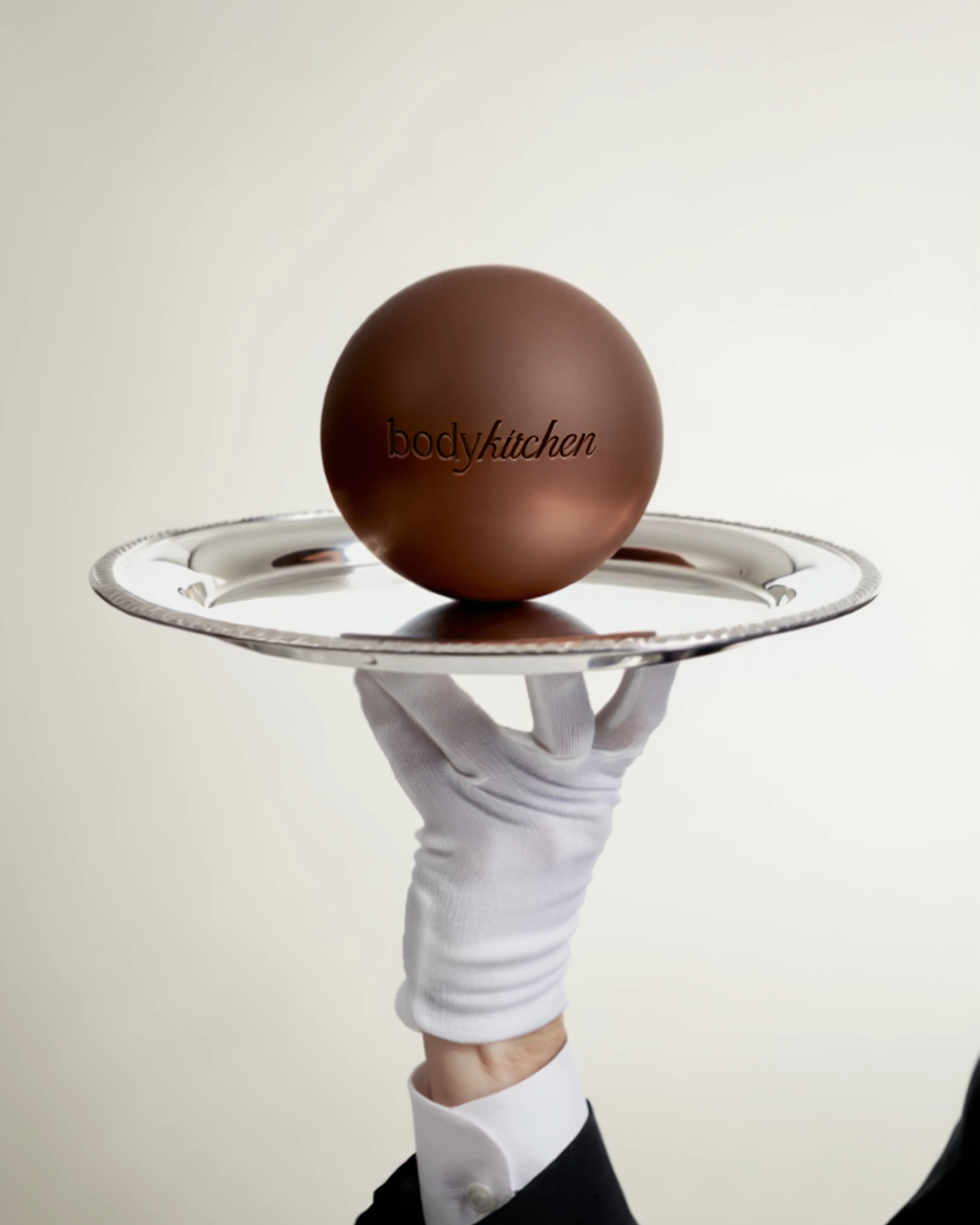 White-gloved hand presenting a BodyKitchen chocolate on a silver tray.
