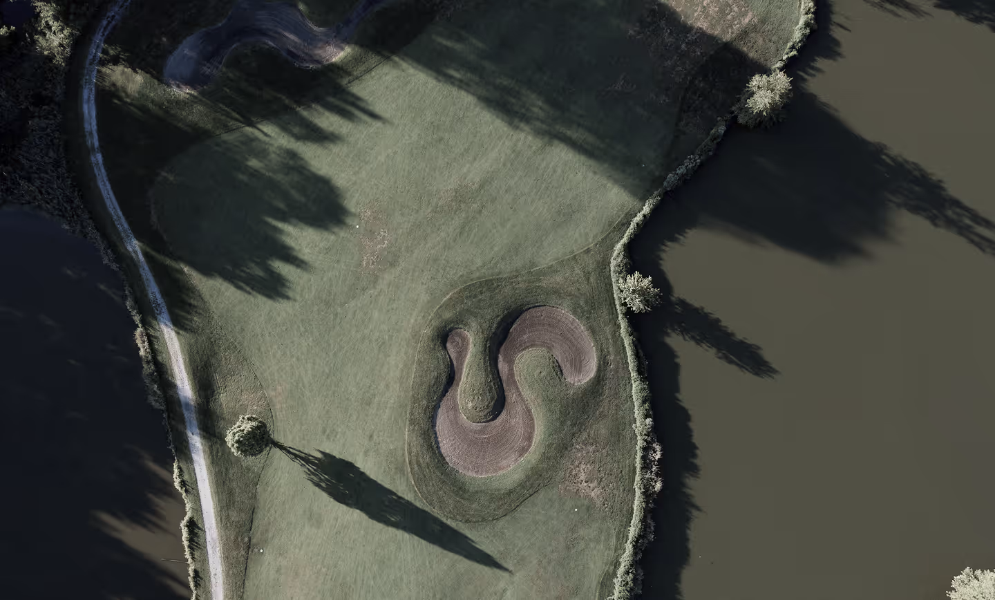 Aerial view of a golf course green bordered by water, featuring an S-shaped sand bunker and surrounding trees casting long shadows.
