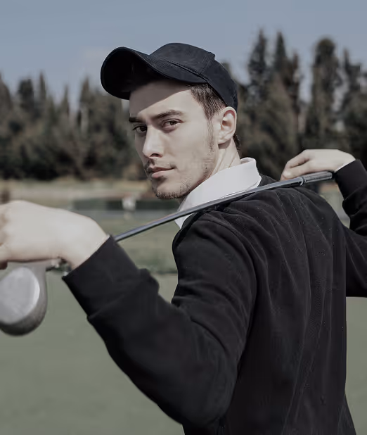 Man wearing a black cap and jacket holding a golf club behind his shoulders on a golf course.