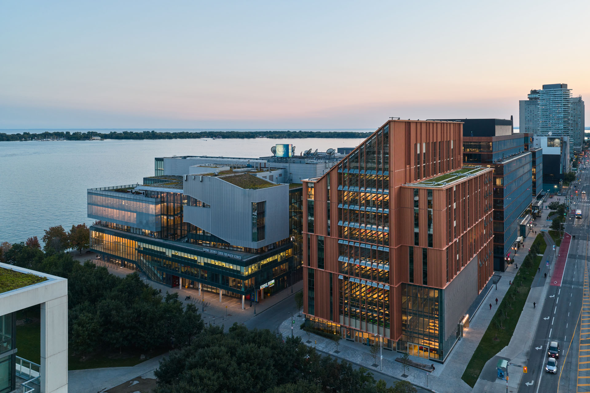 Aerial view of modern buildings near a waterfront at dusk, with lit windows and a street running alongside.