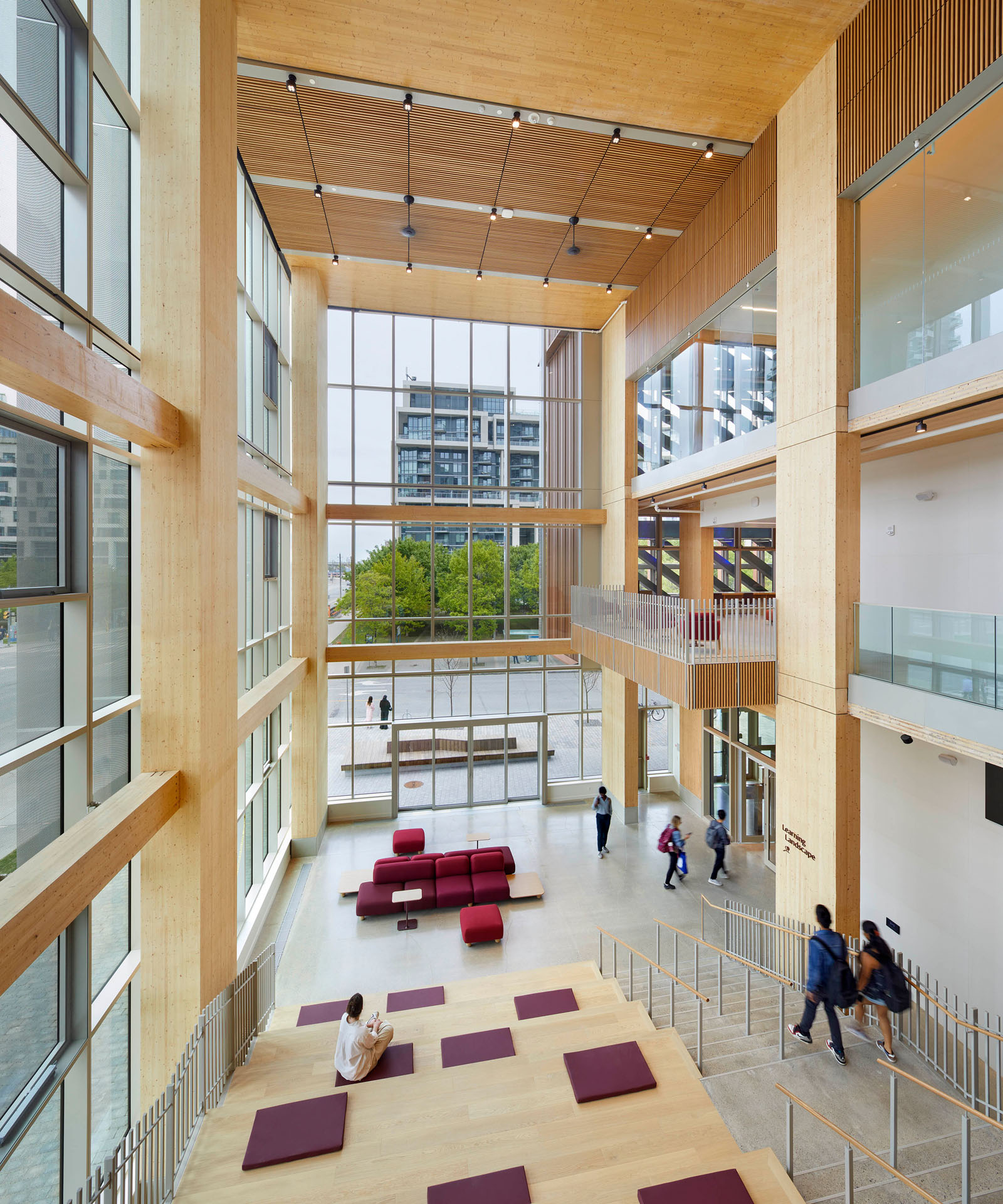 Spacious modern atrium with tall windows, wooden beams, tiered seating with cushions, and several people walking and sitting.