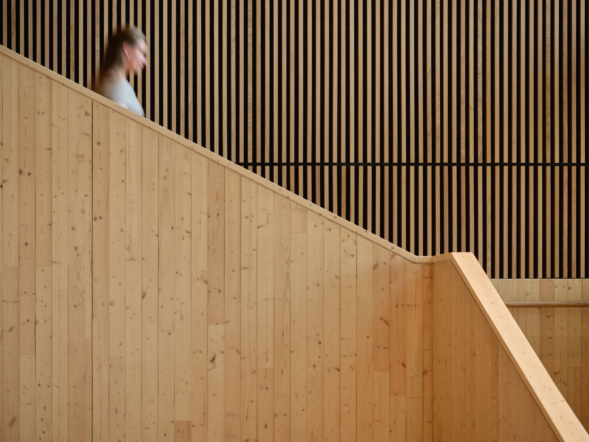 A blurred person walking down a wooden staircase with vertical wooden slats on the wall in the background.