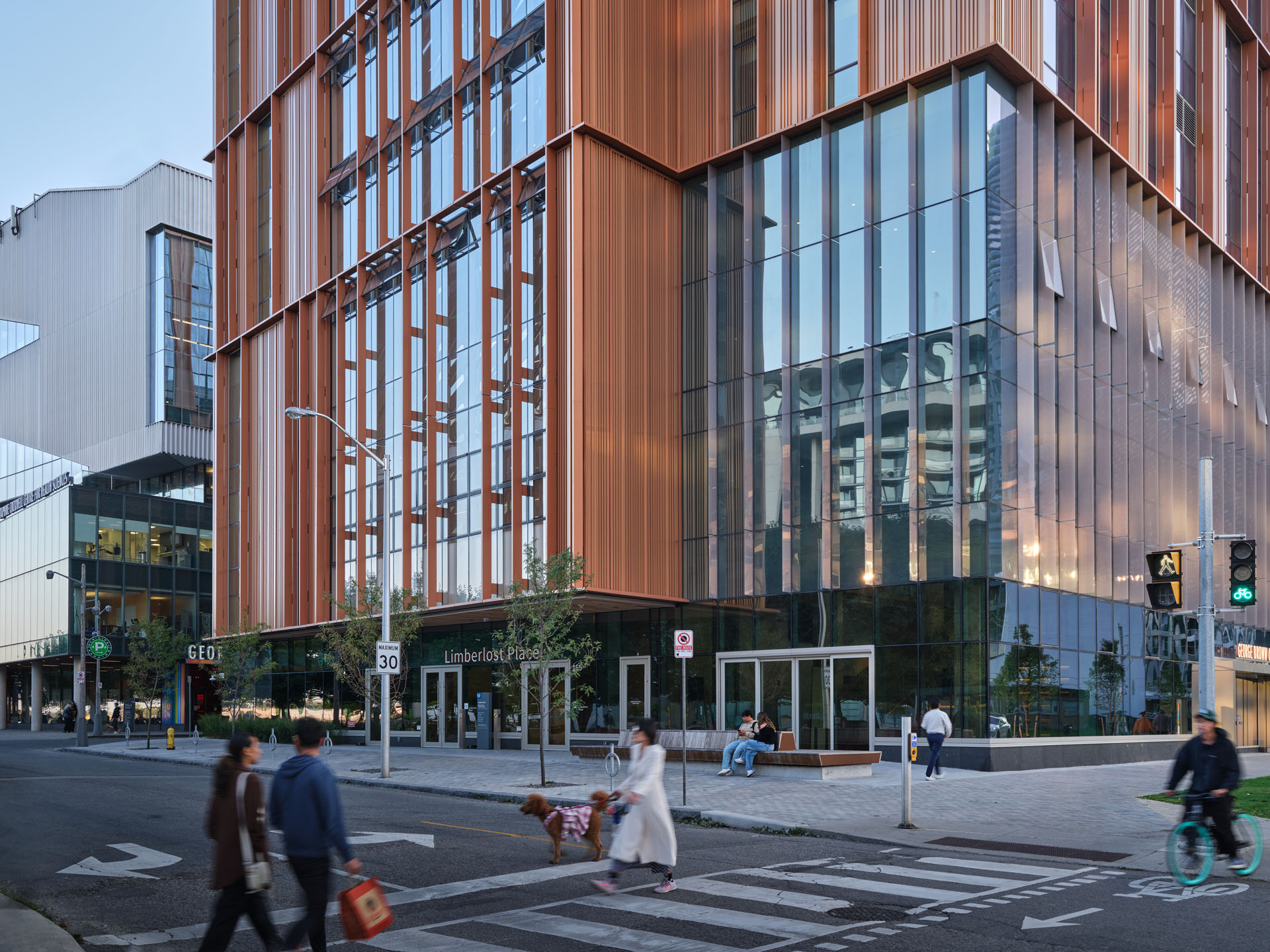 Modern building with large glass windows and vertical orange panels on an urban street with pedestrians, a dog, and a cyclist.