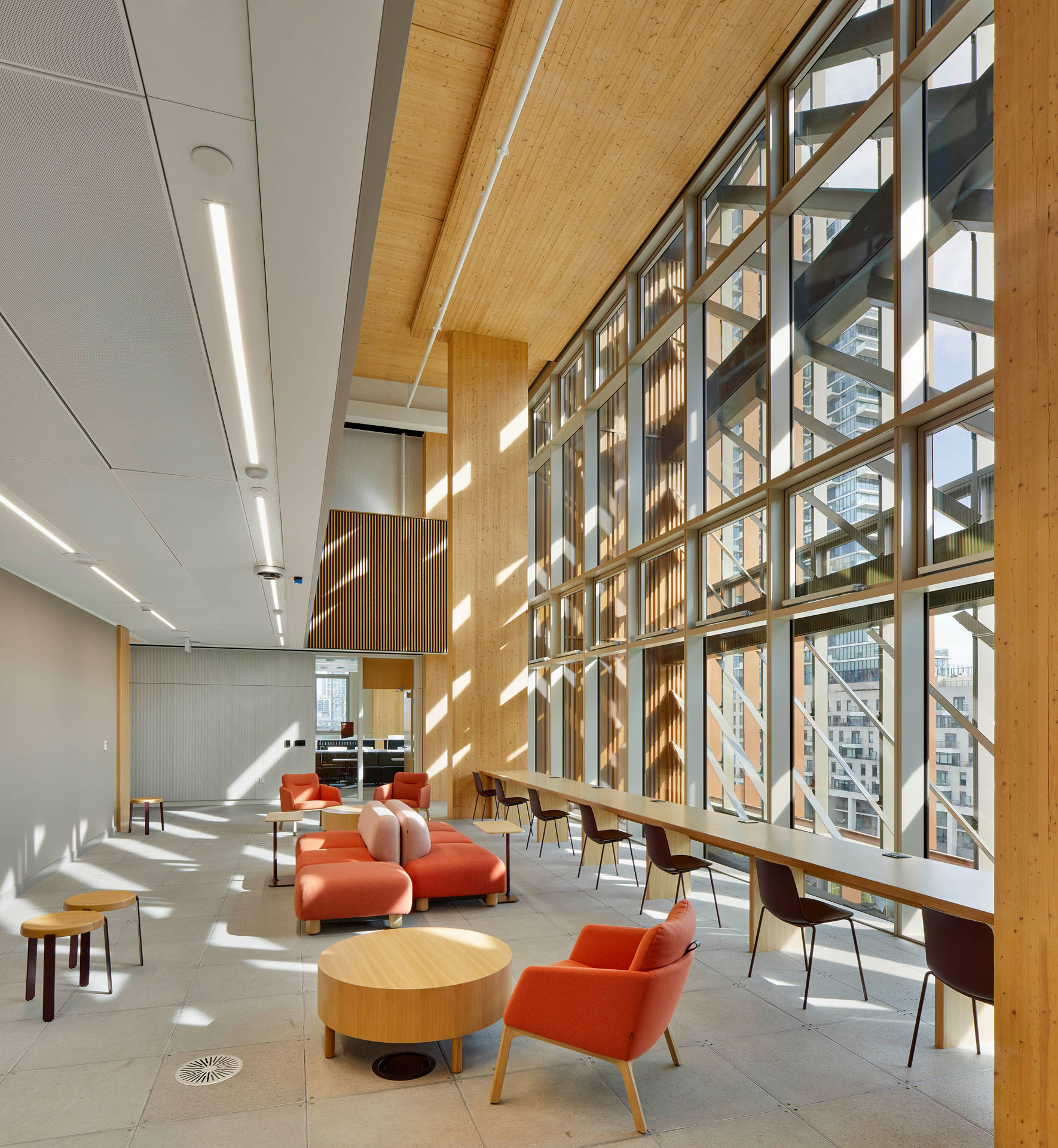 Modern lounge area with large floor-to-ceiling windows, orange cushioned chairs and sofas, wooden tables, and natural wood ceiling.