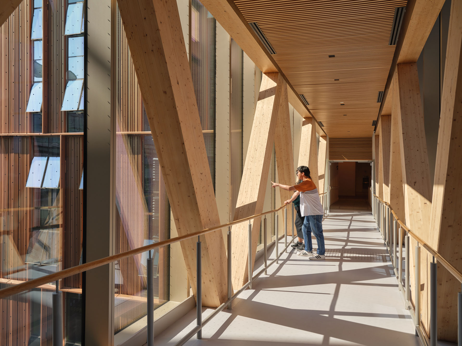 Two people standing on a sunlit corridor with large wooden beams and floor-to-ceiling windows, one person pointing outside.