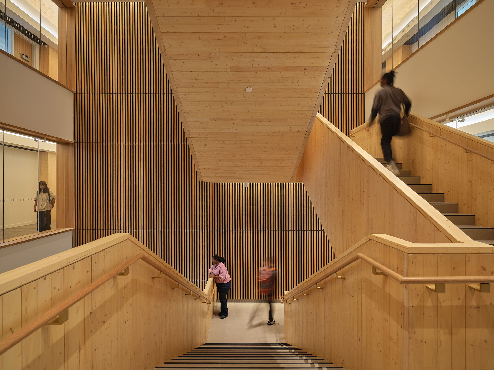 Interior view of a wooden staircase with vertical slatted wood paneling and three people; one leaning on the railing, one walking down, and one ascending the stairs.