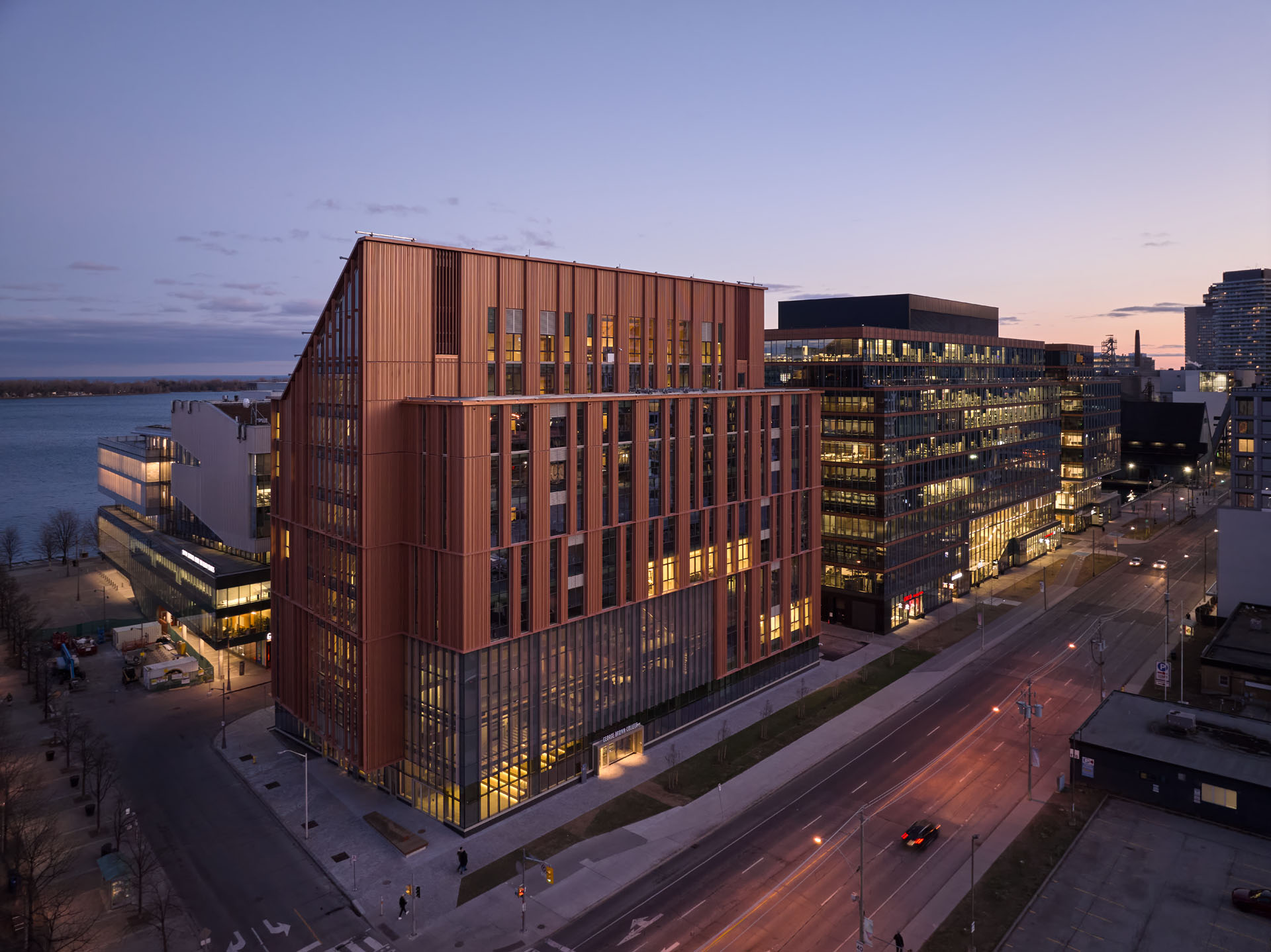 Dusk aerial view of modern office buildings along a waterfront street with illuminated windows and light traffic.