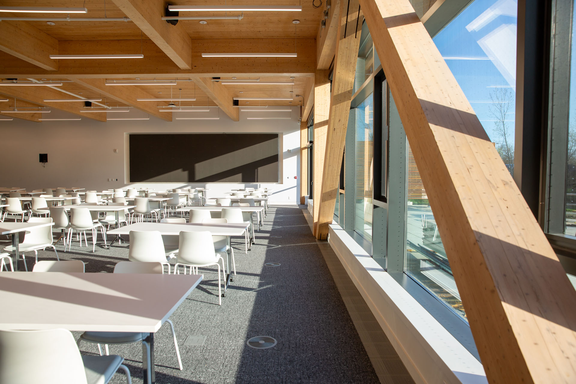 Empty modern classroom with white chairs and tables, wooden ceiling beams, and large windows letting in natural light.