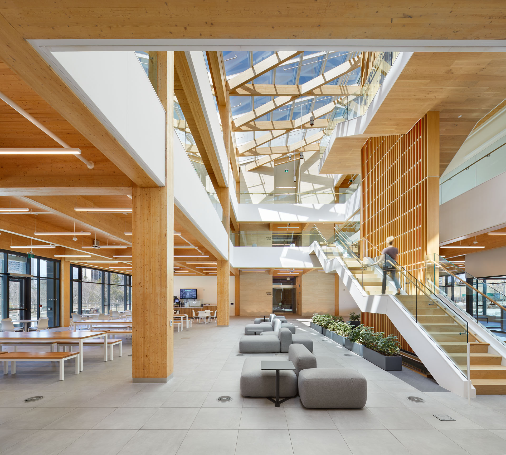 Bright modern open interior with wooden beams, gray modular seating, indoor plants, large windows, and a staircase with glass railings leading to upper floors.