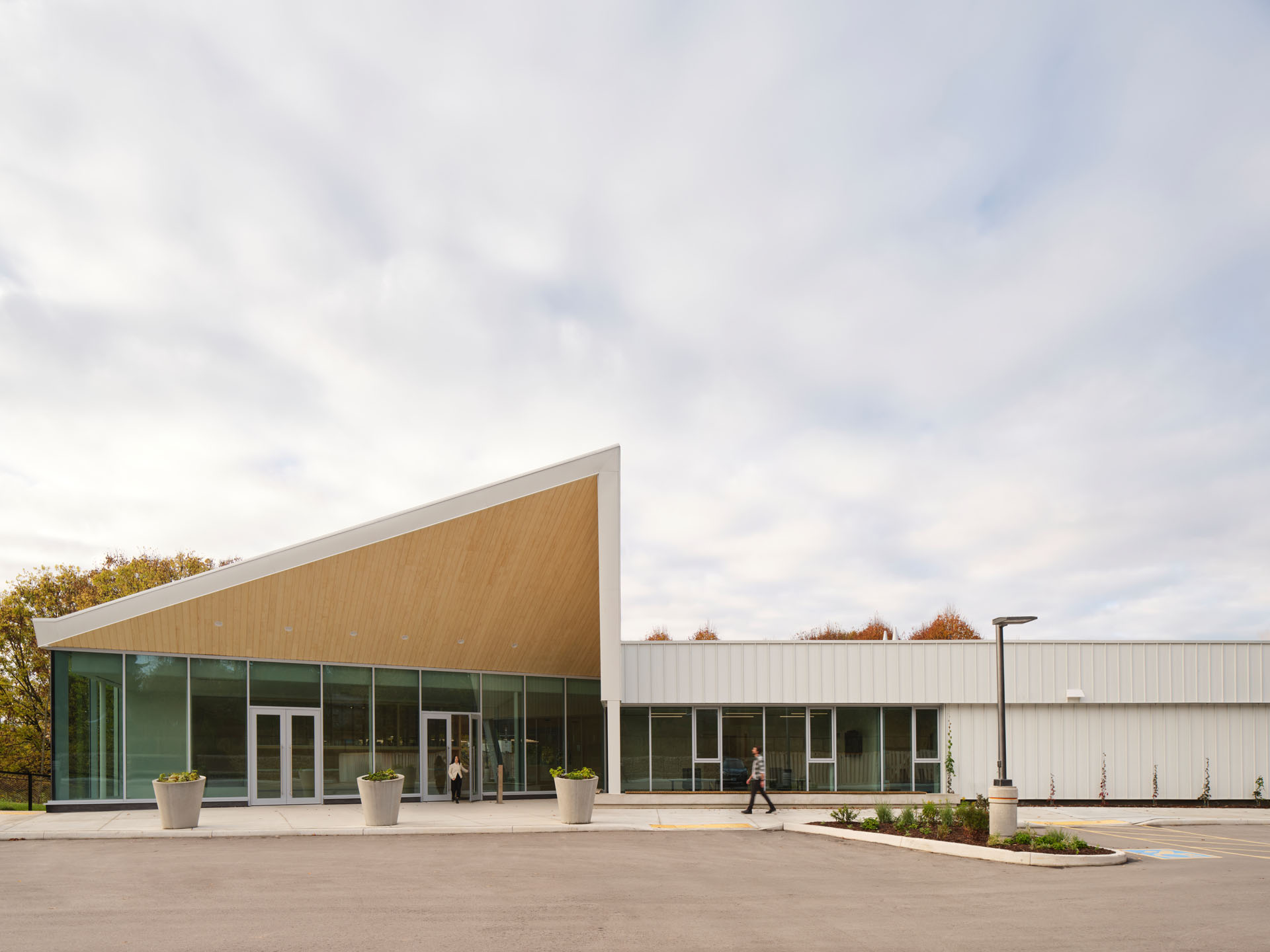 Modern white building with large glass windows and a slanted wooden overhang at the entrance with two people walking outside.