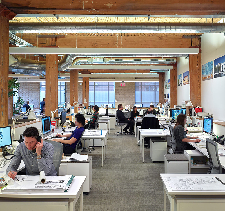 Modern open office with exposed wooden beams and ductwork, featuring employees working at desks with computers and architectural blueprints.