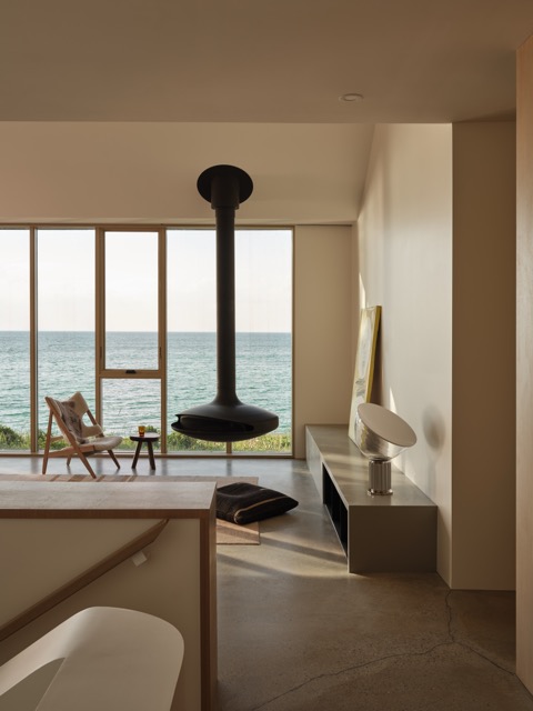 Modern minimalist living room with a hanging black fireplace, wooden chair, small table, and large windows overlooking the ocean.