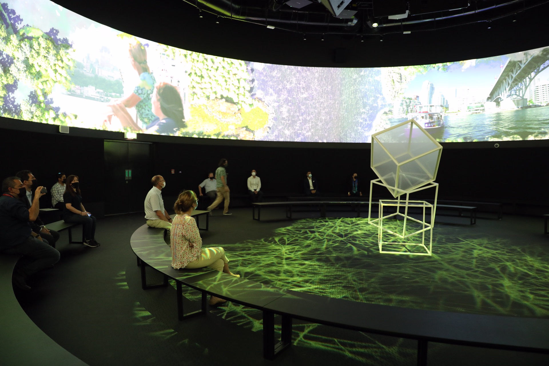 People seated on curved benches in a dimly lit circular room with a cube sculpture in the center and a panoramic video projection around the upper walls.