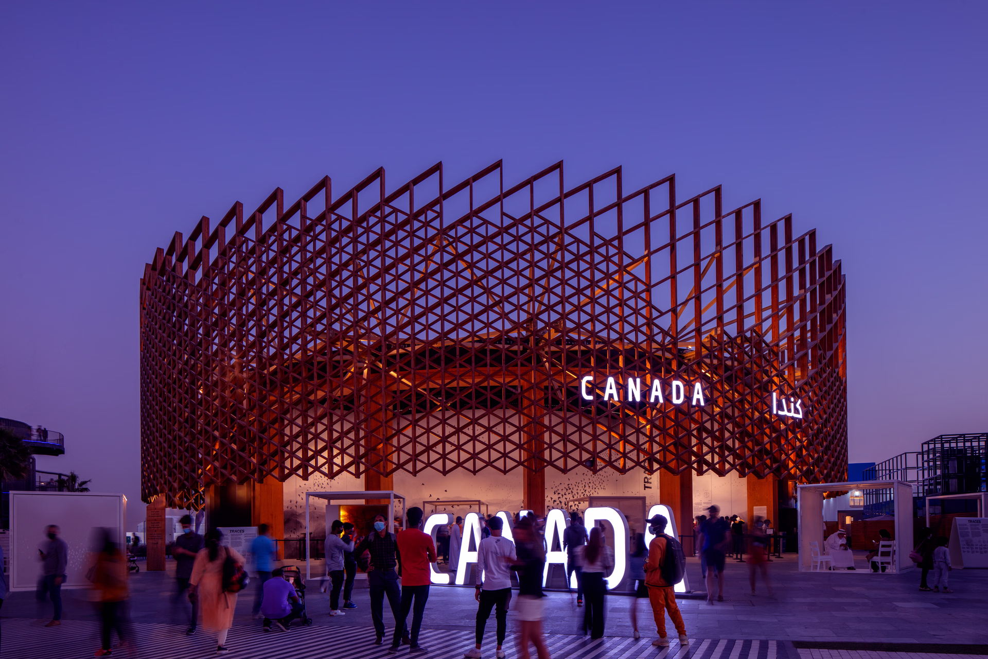 People gathered in front of the Canada pavilion with illuminated Canada signs and a large lattice structure at dusk.