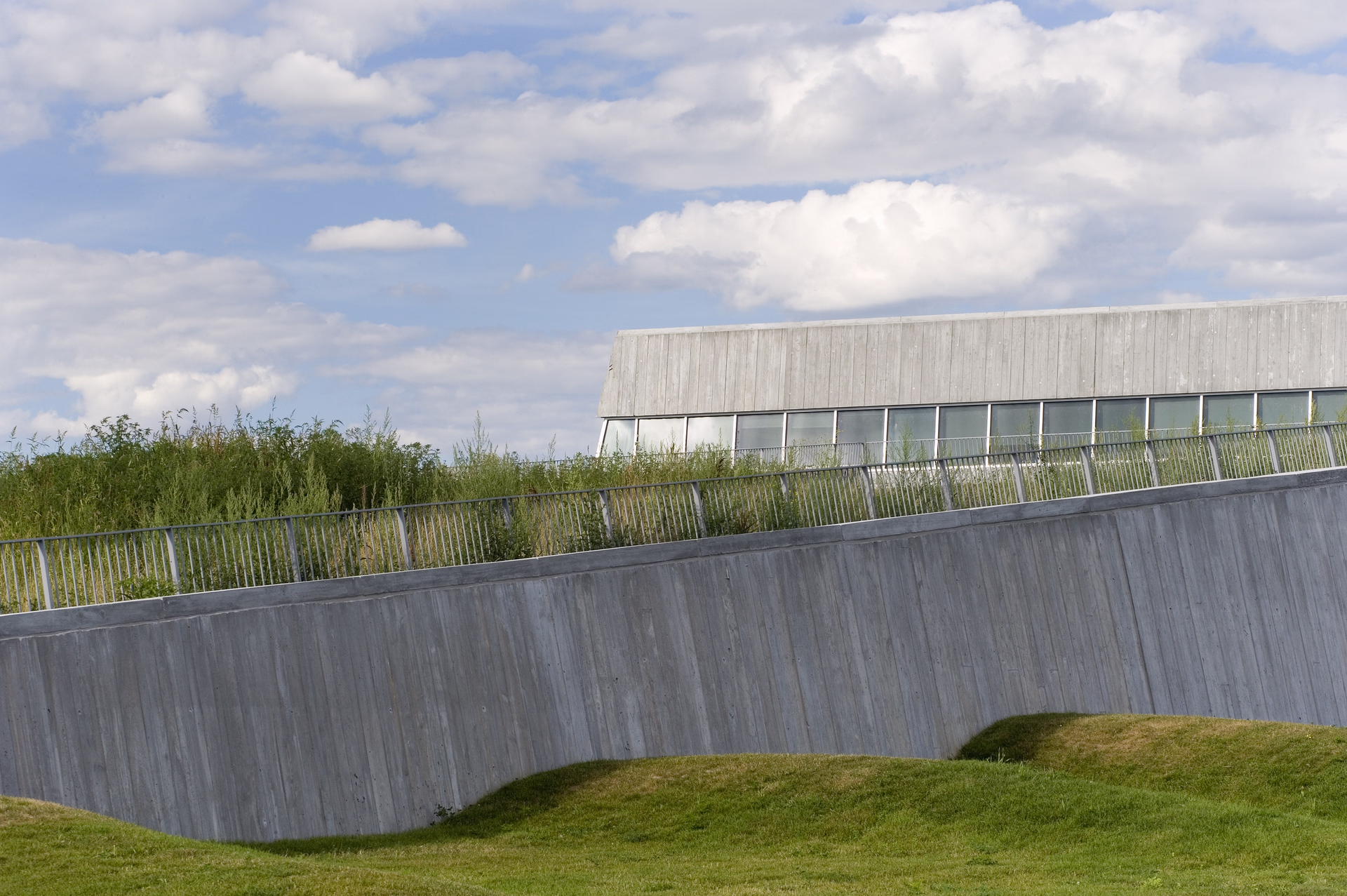 Modern building with slanted concrete wall, a railing, grass, and a cloudy sky.