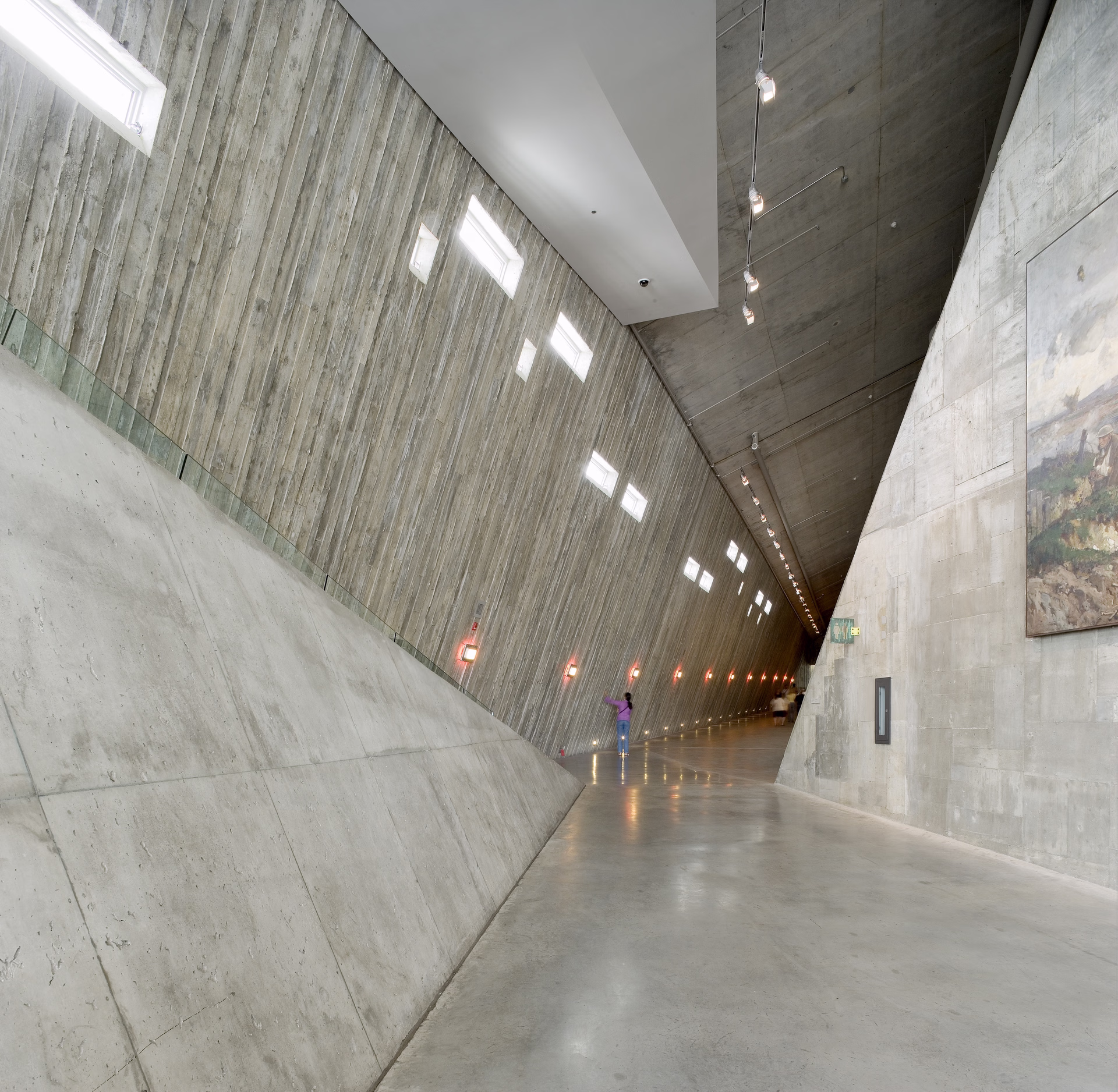 Long, sloped concrete corridor with small high windows and a person standing near the wall.