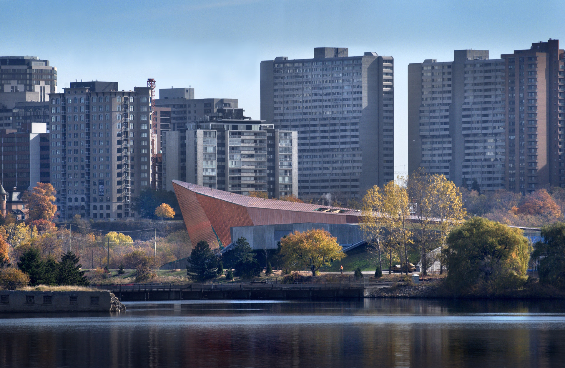 City skyline with tall buildings behind autumn trees and a reflective lake in the foreground.