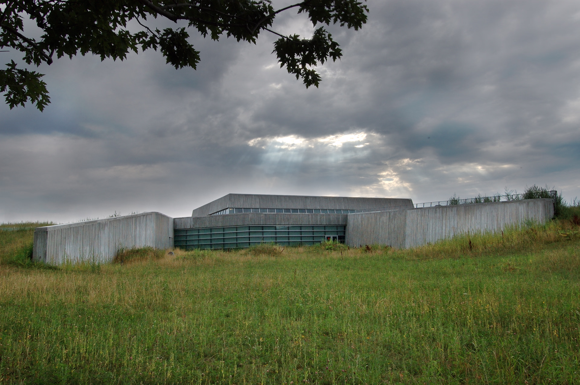 Modern concrete building set in a grassy field under a cloudy sky with sun rays breaking through.