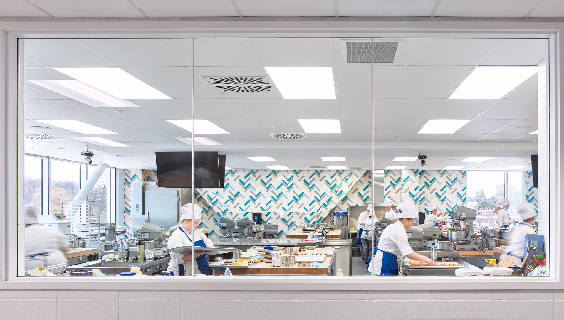 Culinary students in white uniforms and hats working in a bright kitchen classroom with large windows and patterned wall.