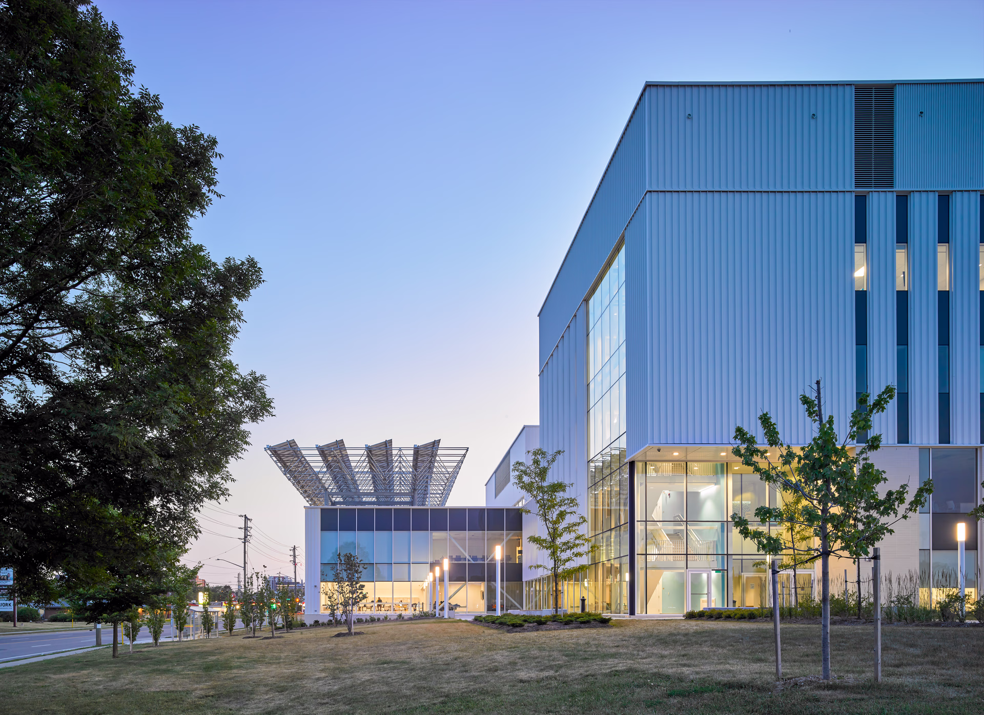 Modern building with glass windows and solar panel structure on the roof at dusk, surrounded by grass and young trees.
