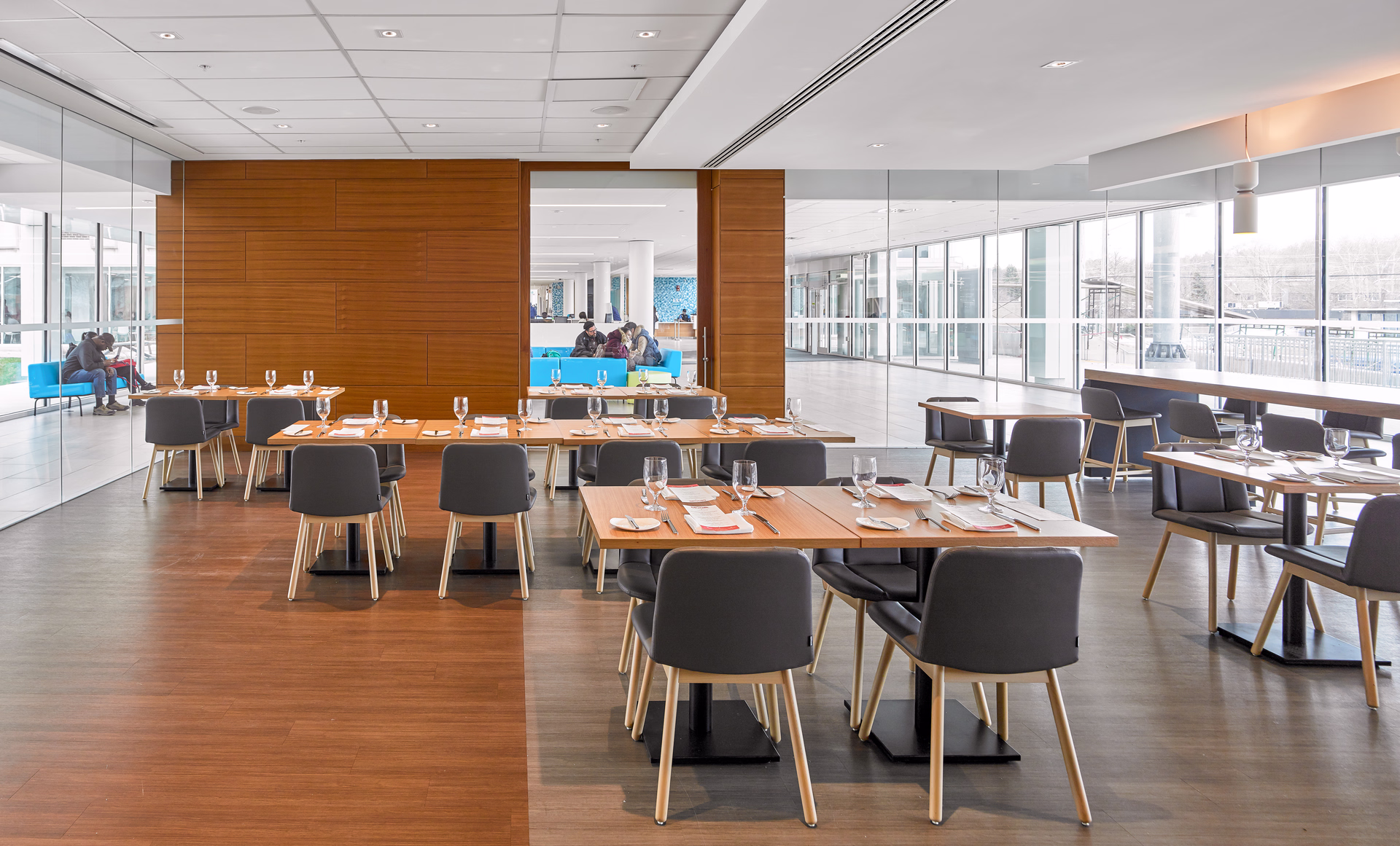 Modern, spacious dining area with wooden tables set with glassware and cutlery, surrounded by gray chairs, and large windows letting in natural light.