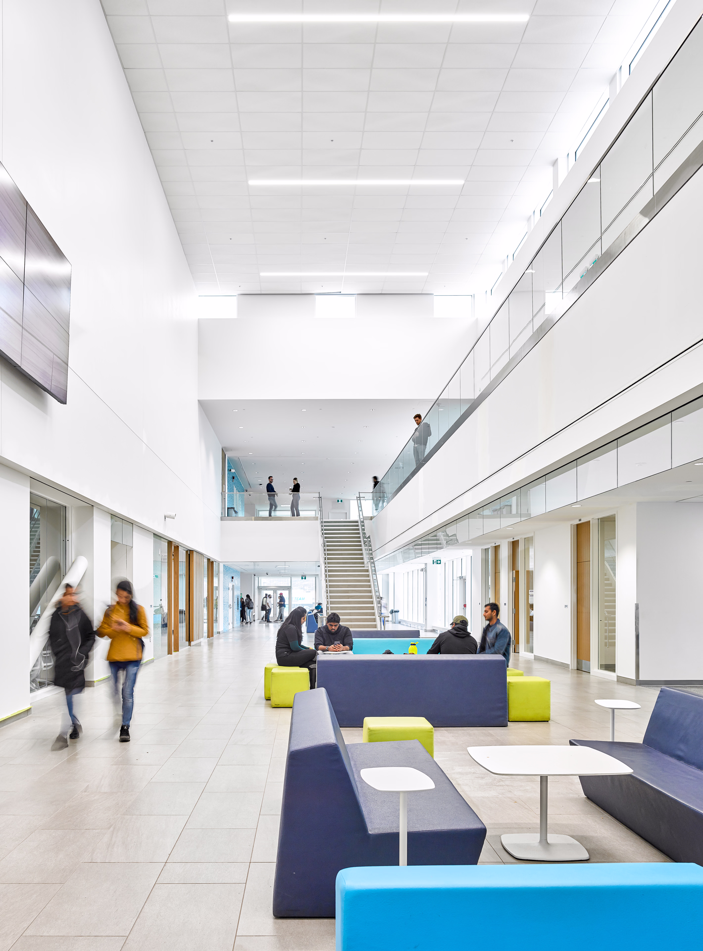 Bright modern lobby with blue and green seating, people sitting and walking, and a central staircase under natural light.