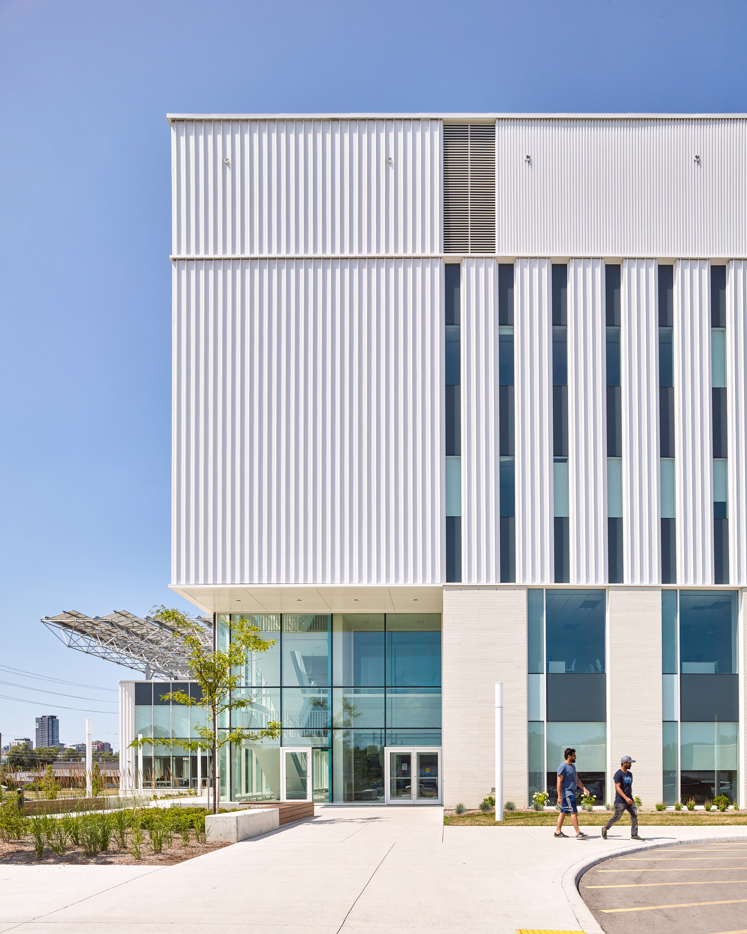 Modern building with vertical white panels and large glass windows under a clear blue sky, with two people walking on the sidewalk.