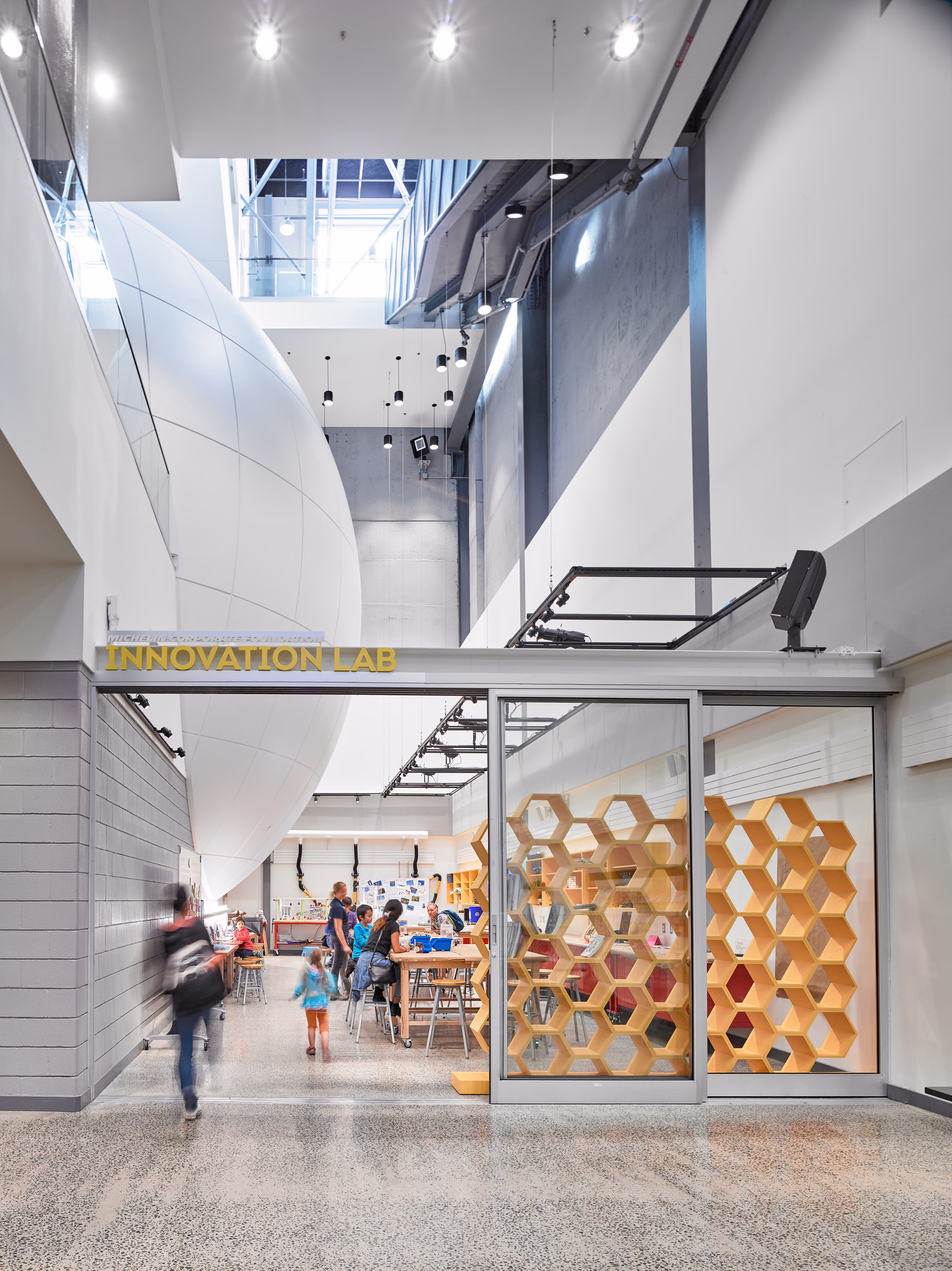 Interior view of a modern Innovation Lab with people working at tables and hexagonal wooden shelves visible through glass walls.