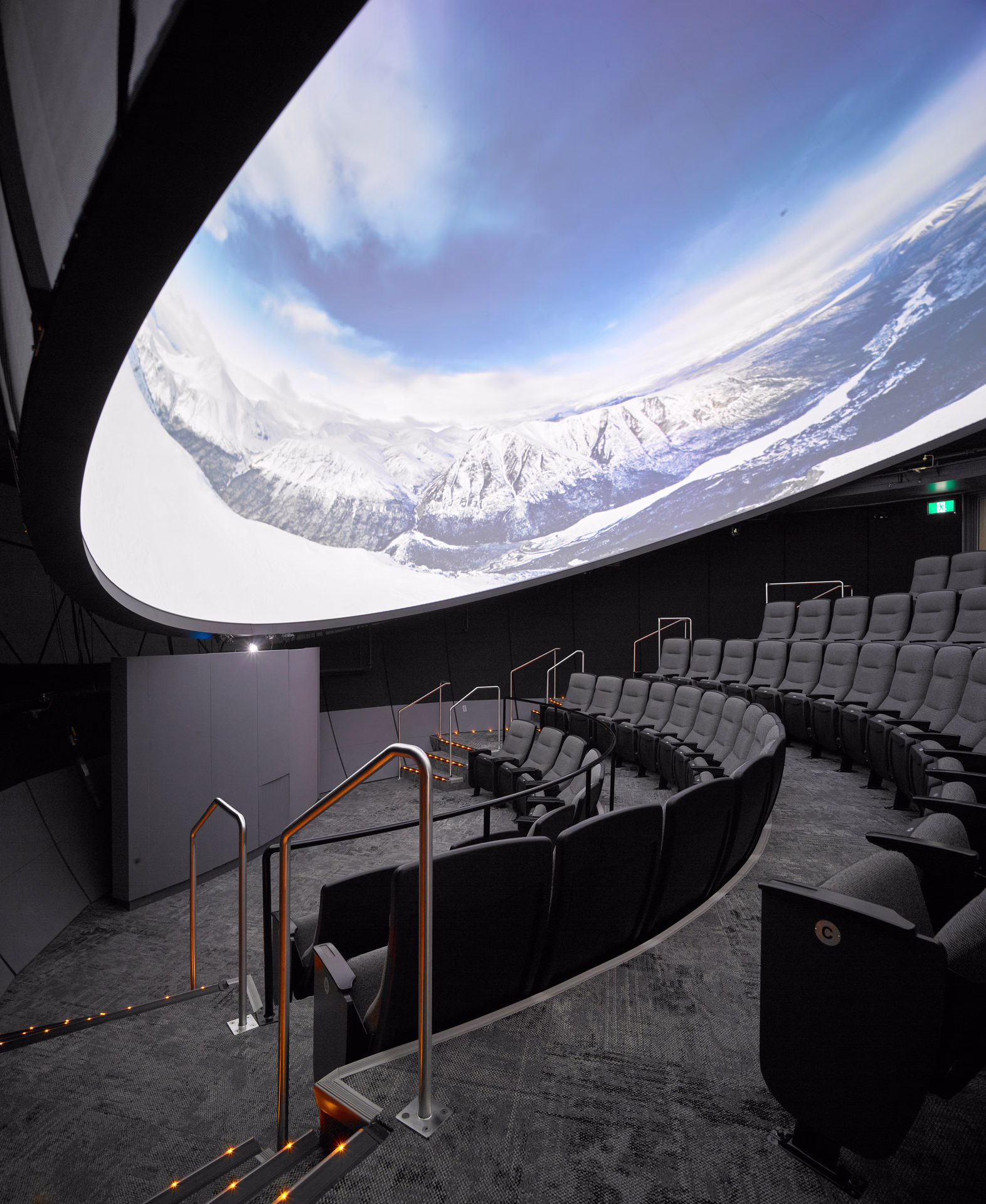Interior of a modern planetarium with curved rows of gray seats facing a large dome screen displaying snowy mountain landscapes under a blue sky.