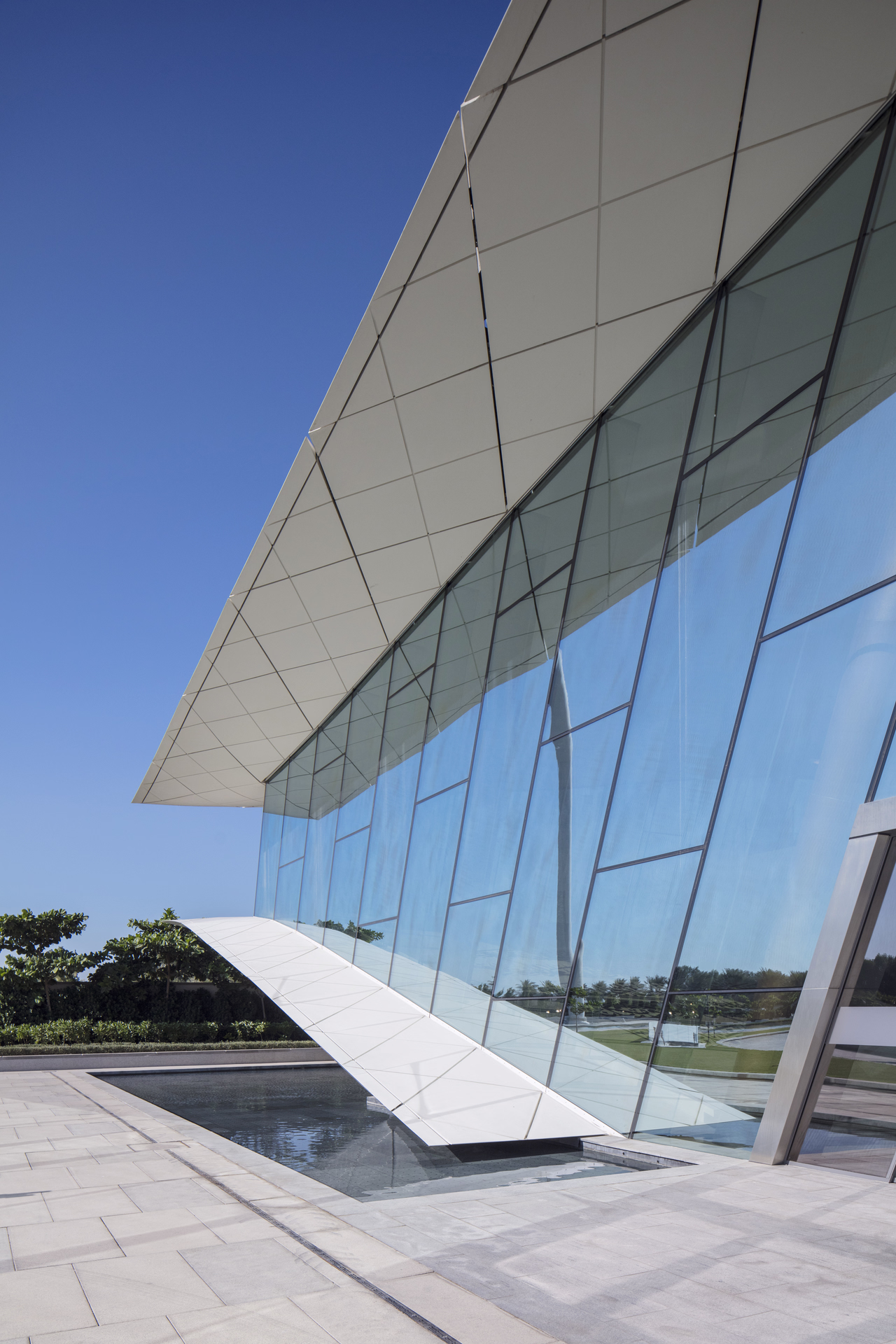Modern building with large angled glass windows reflecting blue sky and surrounding greenery.